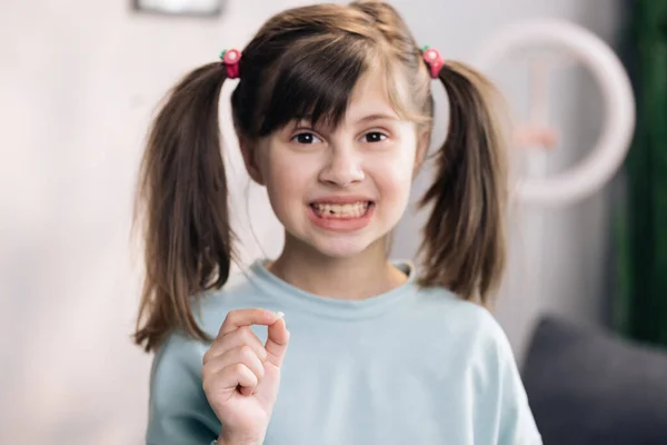 Happy girl holding her fallen tooth in hand. Portrait of cute little child girl is rejoices and showing her lost milk tooth and smiling to camera of toothless mouth while she standing in bedroom — Stock Photo, Image