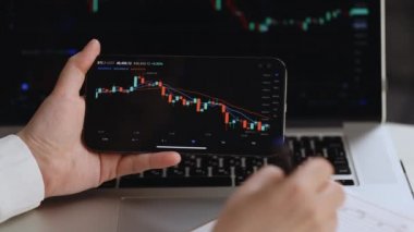 Hisse senedi işlemcisini kapatıyoruz. Not defterine yazıyoruz. Hisse senedi fiyat verileri eski ve yeni telefon üzerinde gösteriliyor. Borsacı ekrandaki verilere bakıyor. Finansal analist