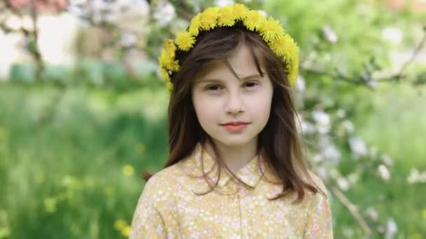 Kafasında çelenk olan küçük kız kameraya bakıyor. Komik Düşünceli Çocuk 'la yüzleş. Yakından gülümseyen bir yüz ve gün ışığında taze bir bahar çiçeği. Çiçek tomurcuklarından hoşlanan mutlu kızın yüzü — Stok video