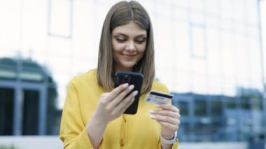Beyaz bir kadın, şehir merkezindeki binalarda cep telefonuyla online alışveriş yapmak için kredi kartı numarasını giriyor. Smartphone iletişim teknolojisi para bankacılık finans broşür ödeme