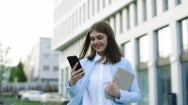 Resmi bir takım elbise giyen beyaz bir kadın cep telefonu kullanıyor. Mesaj yazıyor. Elinde tablet bilgisayarla iş yeri telefonunun önünde yürüyor.