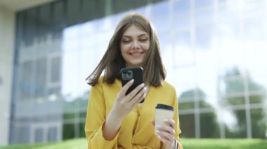 Neşeli genç Avrupalı çekici iş kızı cep telefonu kullanıyor, SMS okurken gülümsüyor, kahve içiyor, dışarıda vakit geçiriyor. Şehir insanlarının portreleri. 4k