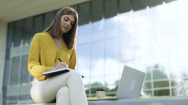 Webinar ya da sanal eğitim uzaktan kumandalı kursları izleyen kız dışarıda, kampüs dışında oturuyor. Beyaz kadın üniversite öğrencisi. İnternette öğreniyor. Laptop kullanıyor, not alıyor..