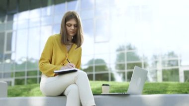 Genç beyaz kadın üniversite öğrencisi dizüstü bilgisayarla öğreniyor, not alıyor, Webinar ya da sanal eğitim uzaktan kumandalı sınıfını izliyor kampüs dışında oturarak eğitim görüyor..