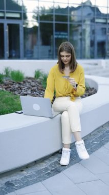 Elinde kredi kartı ve akıllı telefon olan bir kızın dışarıdaki bankta oturduğu dikey video. Çevrimiçi dükkanda hızlı ve kolay ödemeler yapan bir bayan müşteri. E-bankacılık uygulaması