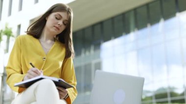 Genç kız öğrenci bankta oturur, uzak bir sınıfı internetten alır, genç kız öğrenci evden ders çalışır, bilgisayardan canlı ders izler, notlar alır, uzaktan eğitim kavramı edinir.