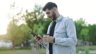 Gün ışığında yakışıklı, beyaz, genç, sakallı bir adam gün batımında telefon kulübesi kullanıyor. Mobil teknoloji. Modern moda iş adamı hücresi. Portre. Açık havada. Yavaş çekim