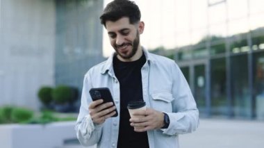 Gülümseyen genç, sakallı bir adam yürüyor. Şehir merkezine bakmak için akıllı telefon kullanıyor. Yakışıklı adam mesajlaşmacı stili. İstihdam teknolojisi. 4k Ağır çekim