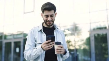 Güzel saç stili ve sakallı çekici sakallı yakışıklı adam akıllı telefonunu kullanıyor ve kahve içerek mesaj yazıyor. Çok şık ve muhteşem görünüyorsun..