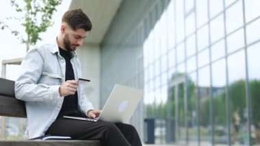 Bilgisayarlı e-bankacılık hizmeti kullanan bir adam güvenli online ödeme yaparak kredi kartını elinde tutuyor. Erkek alıcı internet sitesinde e-ticaret servisi aracılığıyla ödeme yapıyor. Modern ofisin yanındaki bankta oturuyor..