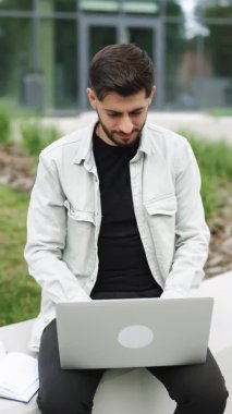 Vertical screen of male professional typing on laptop keyboard. Smiling bearded businessman working on laptop computer near office. Portrait of positive business man looking at laptop screen outdoors