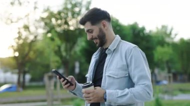 Elinde bir fincan kahve ve modern akıllı telefonla şehir caddesinde yürüyen neşeli genç sakallı adam. Yakışıklı Arap adam iyi haberler okuyor ve gülümsüyor. Yaşam tarzları için teknoloji.