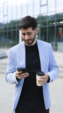 Modern şehir bölgesinde cep telefonlu kahve fincanı kullanan kendine güvenen Ermeni bir adamın dikey ekranı. Pozitif iş adamı açık havada gülümseyen finans haberlerine bakıyor. Başarılı bir yetişkin adam..