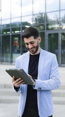 Vertical screen of businessman holding tablet in hands using business apps on tablet computer. Happy face looking tablet screen outdoors. Elegant male chatting online with digital device outside
