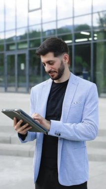Tablet bilgisayardaki iş uygulamalarını kullanarak iş adamlarının tableti elinde tuttuğu dikey format videosu. Mutlu yüz tablet ekranı açık havada. Zarif erkek dışarıda dijital cihazla çevrimiçi sohbet ediyor.