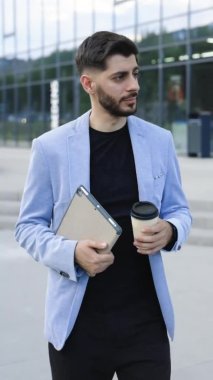 Vertical video of successful young caucasian bearded man with tablet and coffee cup walking between business buildings and smiling with joy. Attractive business male. Career people.