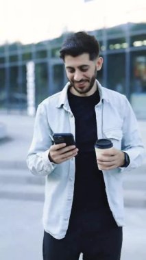 Gülümseyen genç ve sakallı bir adamın portre modu. Şehir merkezine bakmak için akıllı telefon kullanıyor. Yakışıklı adam mesajlaşmacı stili. İstihdam teknolojisi. 4k Ağır çekim