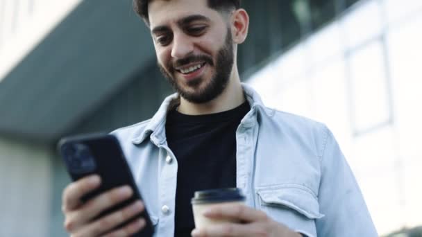 スマートフォンのテキストメッセージにソーシャルメディアアプリケーションを使用して若いエレガントな髭の男は 屋外で笑顔ニュースを受信します 肖像画だ — ストック動画