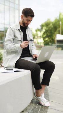 Erkek alıcının modern ofis yakınlarındaki bir internet sitesinden ödeme yaptığı dikey video. Bilgisayarlı e-bankacılık hizmeti kullanan müşteri güvenli online ödeme yaparak kredi kartını elinde tutuyor.