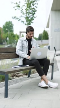İş adamının kulaklık taktığı, bilgisayardan iş görüşmesi yaptığı ve web kamerasıyla iletişim kurduğu dikey bir video. Erkek Yönetici konuşuyor, bilgisayardan sohbet ediyor ofisin yanındaki bankta oturuyor.