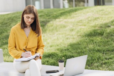 Genç kız öğrenci bankta oturur, uzak bir sınıfı internetten alır, genç kız öğrenci evden ders çalışır, bilgisayardan canlı ders izler, notlar alır, uzaktan eğitim kavramı edinir.
