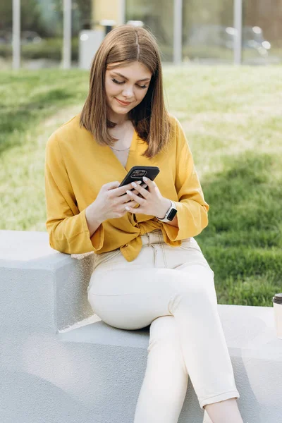 Joyful businesswoman browsing internet on phone, drinking coffee, spending time outdoors. Business woman checking phone content online outdoor. Smiling female freelancer looking phone at remote office