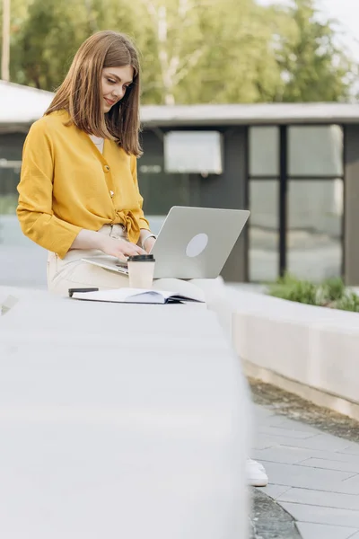 Young european business woman freelancer sitting on bench working with laptop in city park on modern urban street background. Busy worker freelancer working on modern tech notebook device