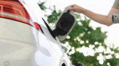 Bottom view of woman hand inserts power cable supply to charge electric or EV car on the green background of blue sky. Woman plugging an electric car or EV at electric charging station.