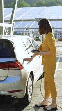 Beyaz tenli bir kadının elektrik prizini açıp şarj istasyonuna şarj ettiği dikey ekran. Güneş panelleri arka planında modern sıfır emisyon otomobilini şarj eden kadın.