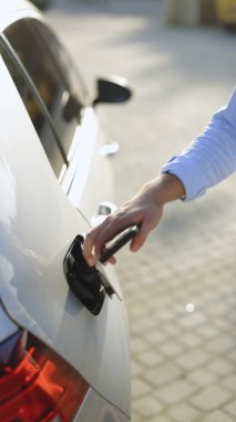 Akıllı telefondaki uygulamayı kullanarak elektrik kablosuna bağlanan bir erkeğin dikey videosu. Elektrikli arabayı şarj istasyonundan çıkaran adam. İş adamı gün batımında şarj istasyonunda elektrikli araba şarj ediyor.