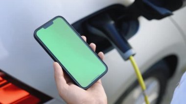 Yeşil ekran, caddede şarj olan elektrikli arabalı akıllı telefonu gösteren kromakey modeli. Cep telefonundaki uygulamaya bakan adam elektrikli aracı kabloyla şarj ediyor..