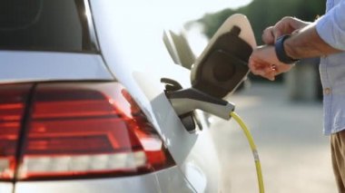 Male hand unplugs power connector into EV car and charges batteries, uses smart watch to disconnect charging. Unrecognizable man disconnects charging cable to to electric vehicle using smartwatch.