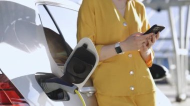 Woman stands near recharging electric car with mobile phone. Vehicle charging at public charging station. Electric car charging near an solar power plant.