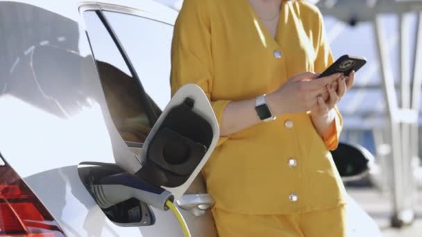 Женщина Стоит Возле Подзарядки Электромобиля Мобильным Телефоном Зарядка Транспортного Средства — стоковое видео