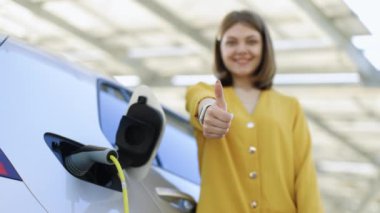 Happy confident young professional business woman leader winner employee looking at camera showing thumbs up hand sign gesture standing on charging station near electric car outdoors.