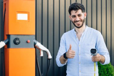 Elektrikli araba şarj istasyonunda elektrik kablosu tutan bir erkek el görülüyor. Sakallı adam elektrik şarj istasyonunun yanında durmuş kameraya bakıyor ve parmağını gösteriyor..
