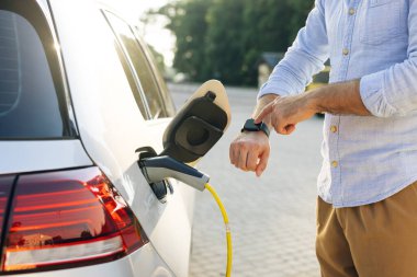 Erkek el güç bağlantısını EV 'ye yerleştirir ve pilleri şarj eder, aktifleştirmek için akıllı bant kullanır. İşadamı elektrikli araca kablo şarj ediyor.
