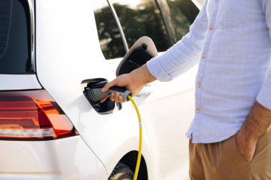 Elektrikli araç şarj istasyonunda güç kaynağı kablosu tutan adam. Şarj cihazının fişini tutan yakışıklı adam şarj istasyonunda şarj olurken....