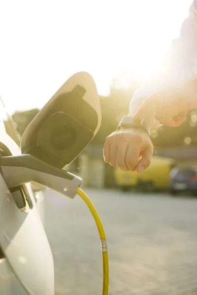 Unrecognizable man plugging in charging cable to to electric vehicle. Male hand inserts power connector into EV car and charges batteries uses smartwatch for activates start charging.