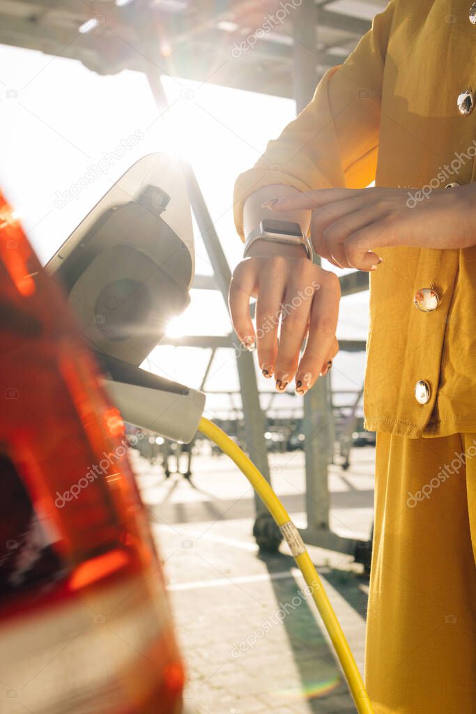 Close up of woman hands attaching power cable supply to charge electric or EV car using app on wearable smart watch. Female plugging an electric car or EV at electric charging station.