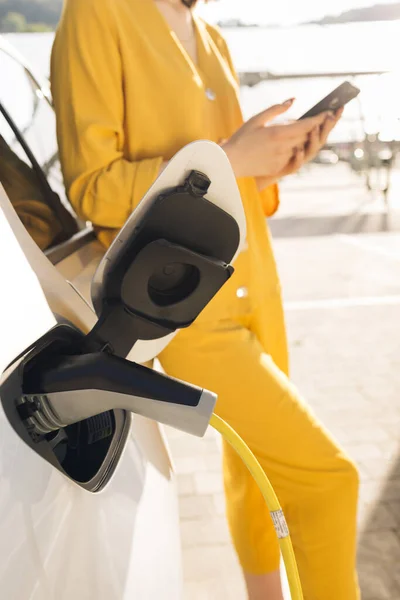 Woman stands with phone near her electric car and waits when vehicle will charged. Female leaning standing near car and use on her phone. Girl waiting while her electric car is charging.