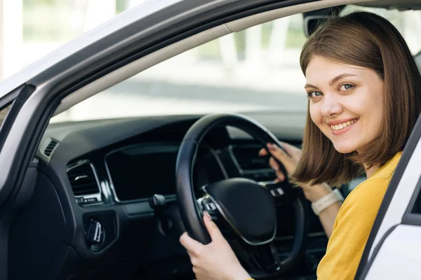 Attractive young business woman looking over her shoulder while driving a car. Driver smiles at camera. Happy millennial woman taxi driver sitting in car holding to steering wheel