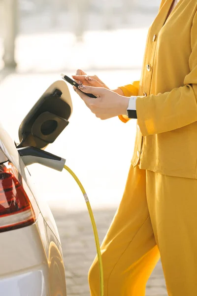 Woman connects an electric car to the charger and adjusts the process of charging the car battery using a cell phone smart phone. Girl plugs power cable to charge electric car in parking lot.