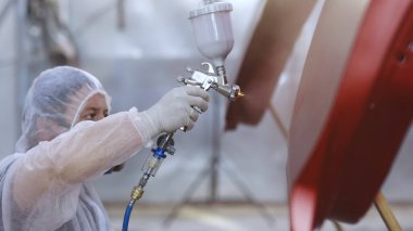 Usta araba servisinde otomobilin bir parçasını boyuyor. Elinde tutarak, koruyucu üniforma giymiş bir şekilde sprey sıkma makinesi kullanıyor. Modern araba boyama