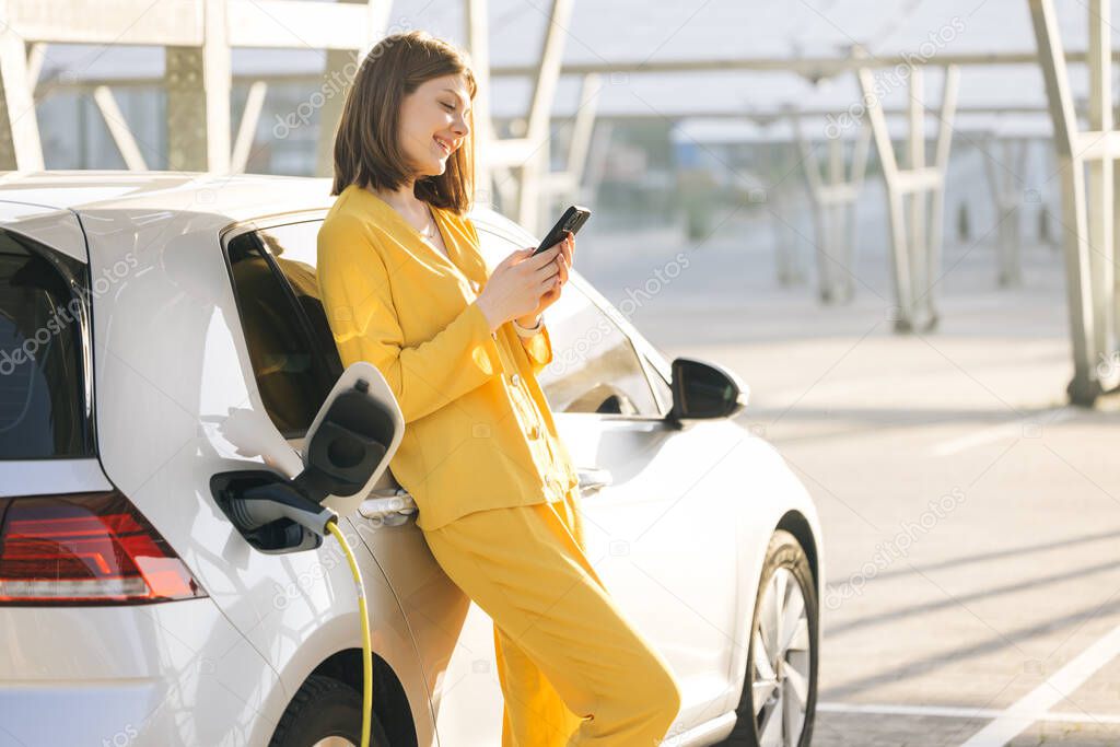 Caucasian woman charging an electric car at a charging station near an electric solar power plant, using her smartphone while waiting.