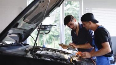 Manager checks tasts on tablet computer and explains an Engine breakdown to an male mechanic. Car service employees inspect cars engine bay with a LED lamp. Modern clean workshop.