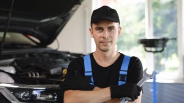Portrait of happy young caucasian man in uniform with tools in his hands working in car service as mechanic, looking at the camera and smiling.