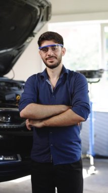 Vertical Portrait Video of Bearded Handsome Car Mechanic is Posing in a Car Service. He Wears a Jeans Shirt and Safety Glasses. His Arms are Crossed. Specialist Manager Looks at a Camera and Smiles.