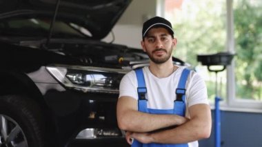 Young caucasian bearded man in blue overalls and black cap looks into camera, while smiling and crosses arms. Male car mechanic at workplace in spacious repair shop.