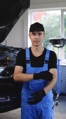 Vertical video portrait of car mechanic crossed arms in car workshop smiling and looking into camera. Concept: repair of machines, fault diagnosis, technical maintenance and on-board computer.